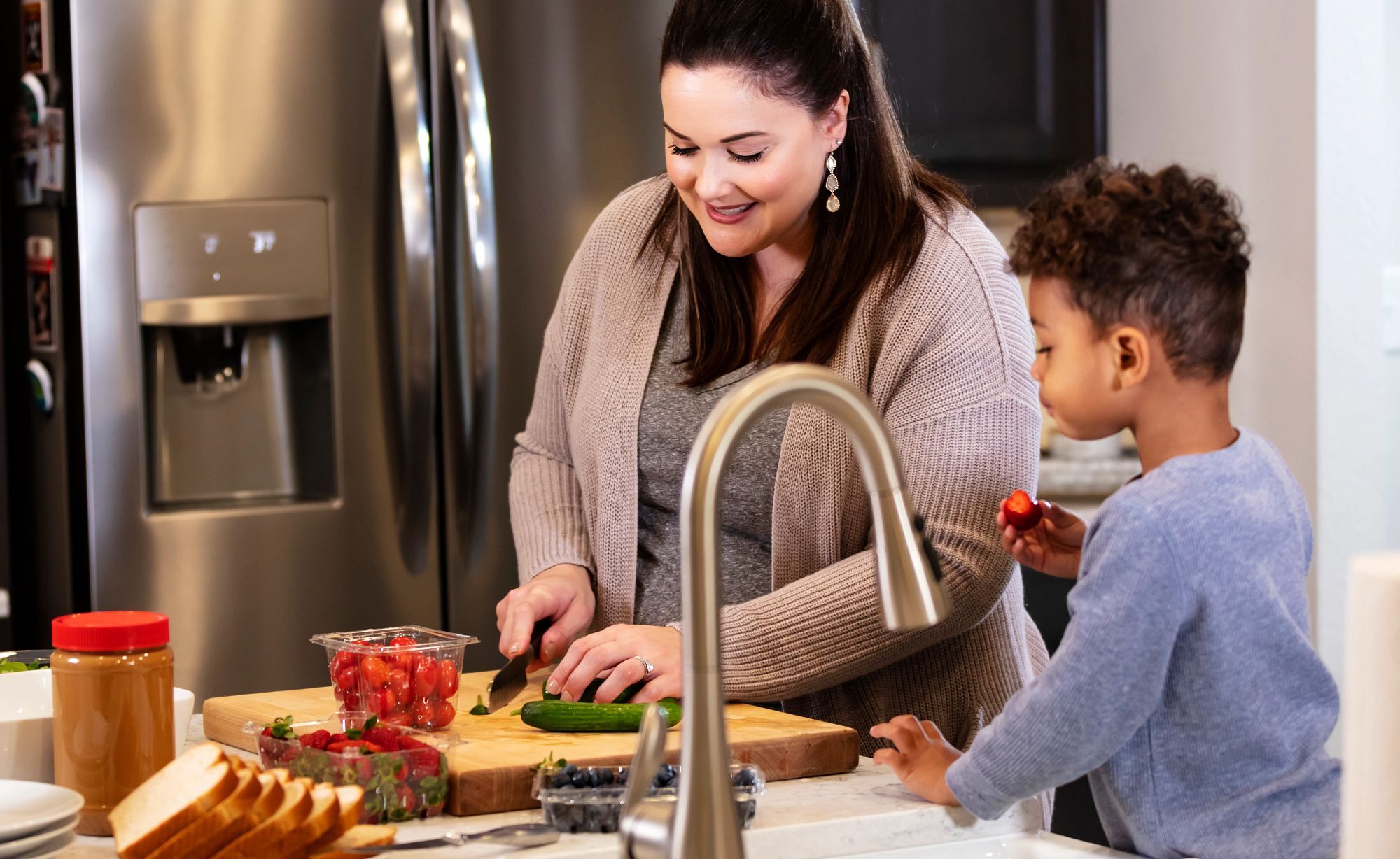 A mother and her son standing in the kitchen as she prepares food for lunch. He is eating a strawberry while he watches. Mom is Caucasian, in her 30s and the boy is mixed race Black and Caucasian, 3 years old. The focus is on the woman.