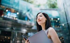 Young Asian businesswoman carrying smartphone and laptop, outside of corporate buildings in the city - Digital Solutions | NSF